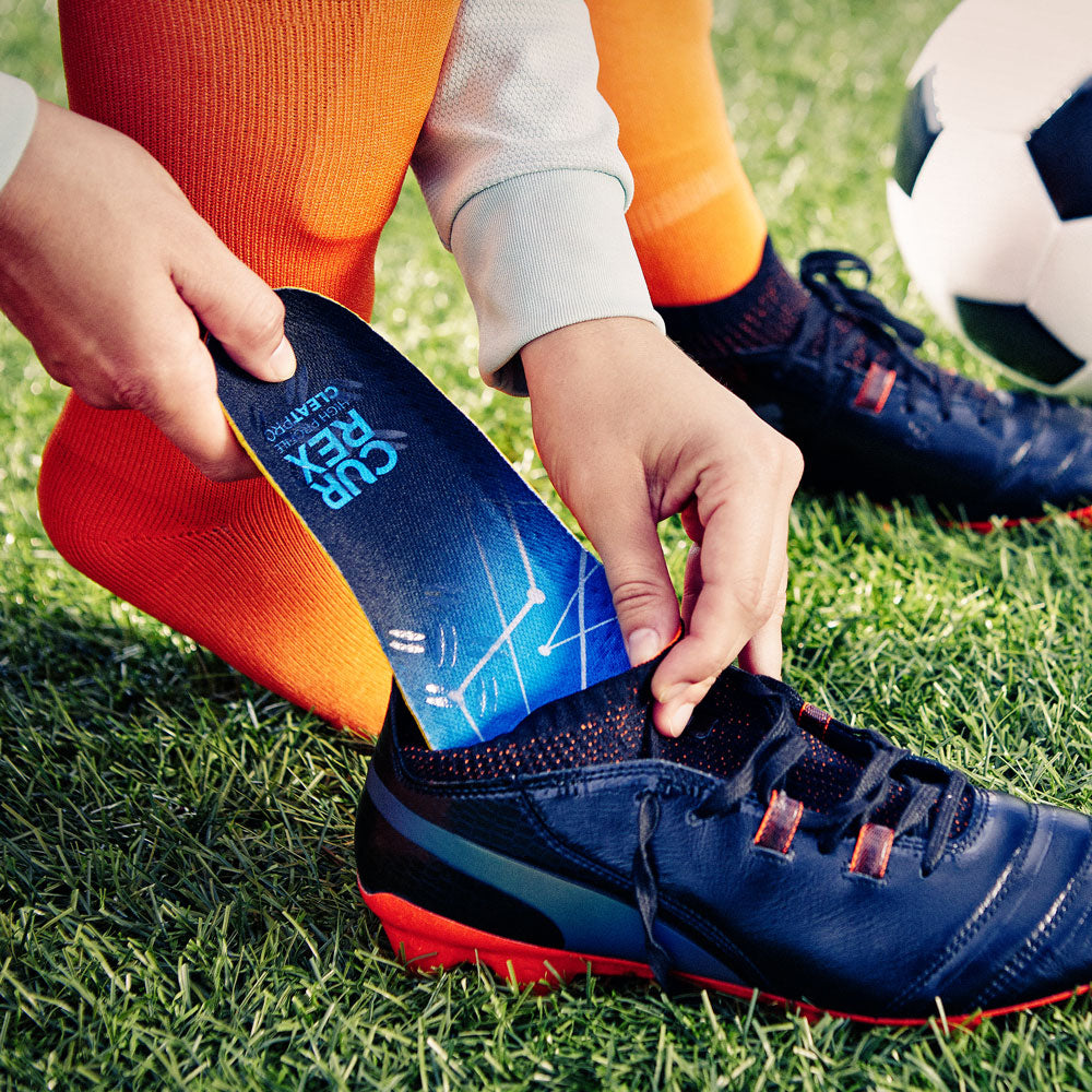 Personne plaçant des chaussettes CURREX CLEATPRO high profile dans des chaussures noires sur un terrain de football #1-choisir-son-profil_med