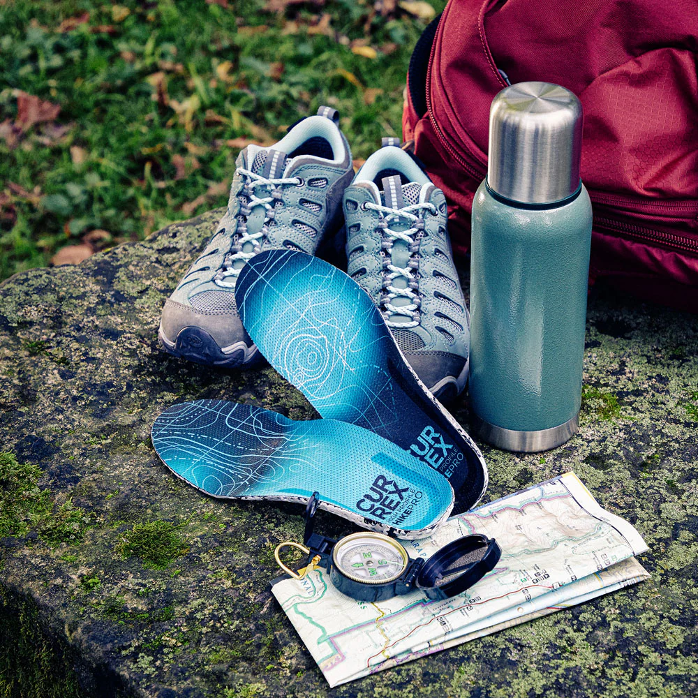 Chaussures CURREX HIKEPRO à côté d'accessoires de randonnée : chaussures, thermos, sac à dos, boussole et carte assis sur une roche recouverte de mousse #1-choisir-son-profil_low